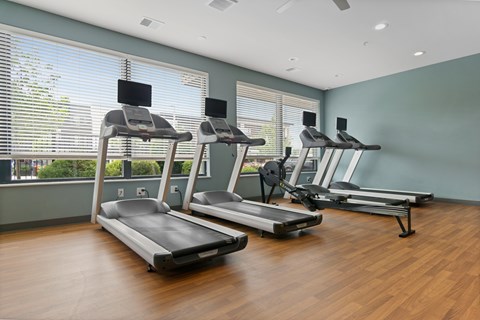 Three treadmills are lined up in a row in a room with wood floors and windows.