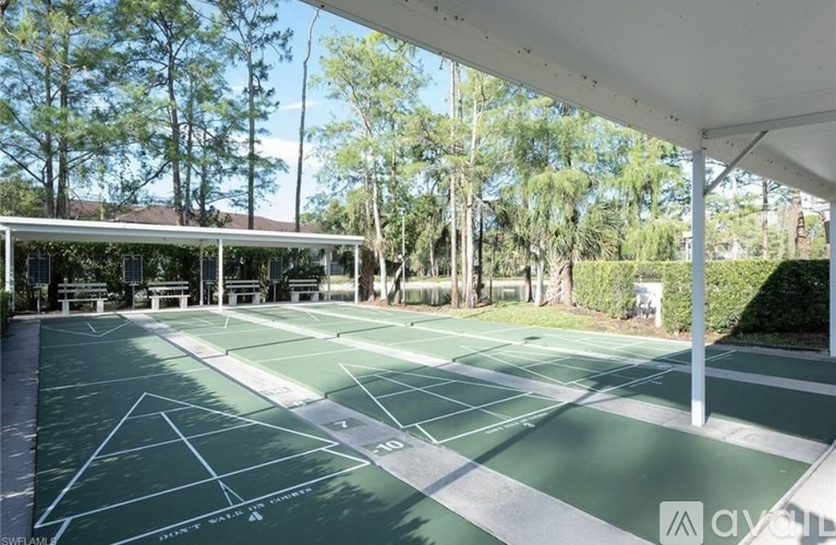 A tennis court with a green surface and white lines, surrounded by trees and a covered area.