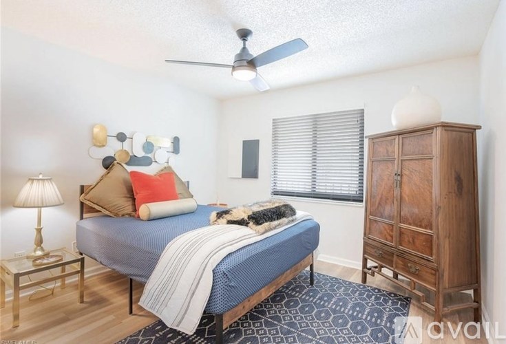 A bedroom with a bed, a nightstand, a lamp, a fan, a window with blinds, and a wooden wardrobe.