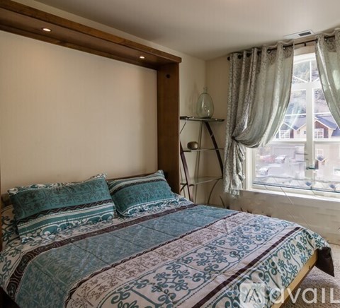 A bed with a blue and white patterned comforter and matching pillows sits in a room with a window and a lamp on a shelf.