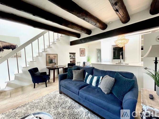 A blue couch with pillows is in the foreground of a well-lit room with a staircase in the background.