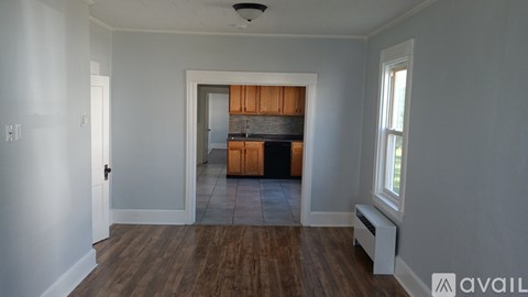 A room with wooden floors and white walls, leading to a kitchen area.