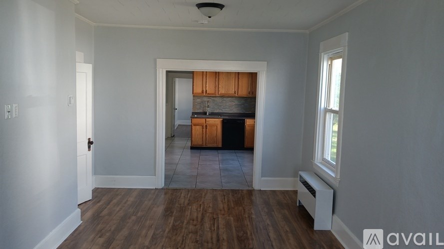 A room with wooden floors and white walls, leading to a kitchen area.