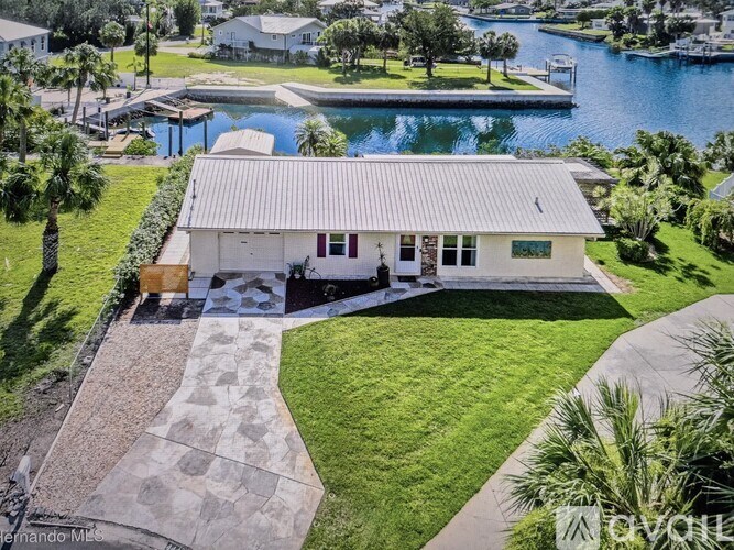 A house with a white exterior and a grey roof is surrounded by a grassy area and a stone pathway leading to a dock.