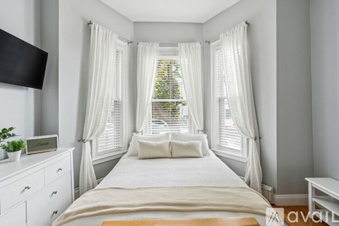 A bedroom with a large bed, a TV on the wall, and a window with white curtains.