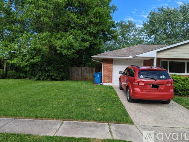 A red car is parked in front of a house with a blue trash can.