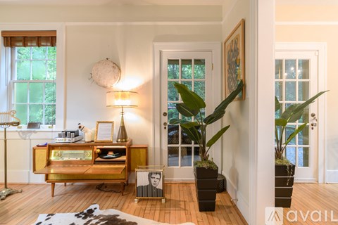 A room with a wooden floor, a white wall, a window with a view of trees, a gold cabinet with a plant on top, and a lamp on the side table.
