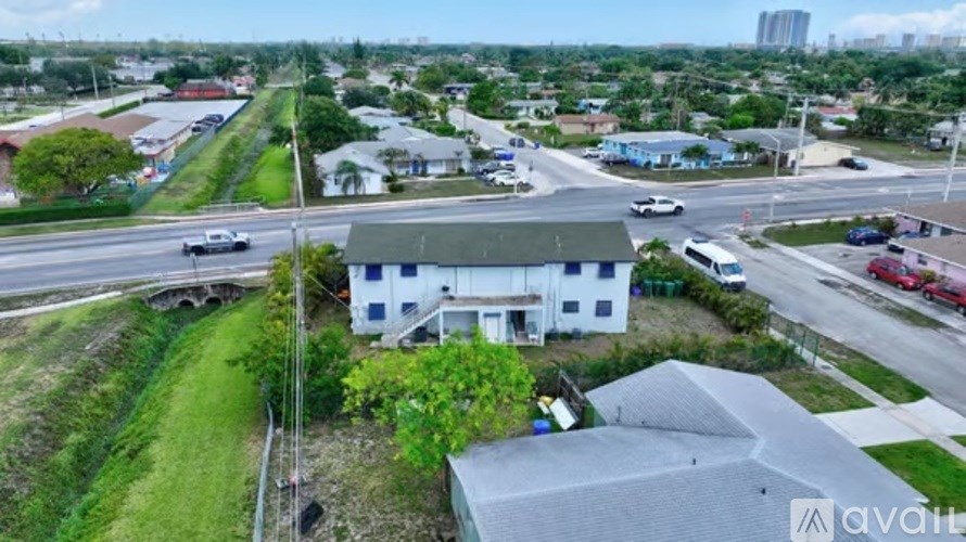 A white building with a grey roof is surrounded by greenery and a road with cars.