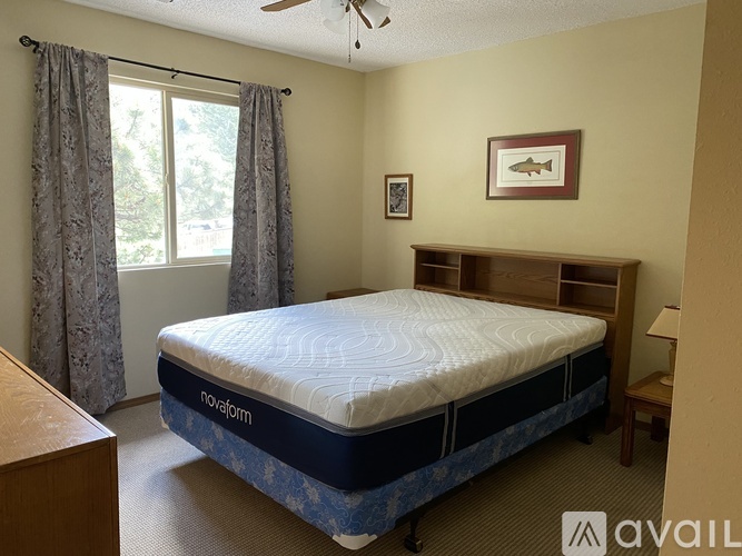 A bedroom with a bed, a window with curtains, a ceiling fan, and a wooden nightstand.