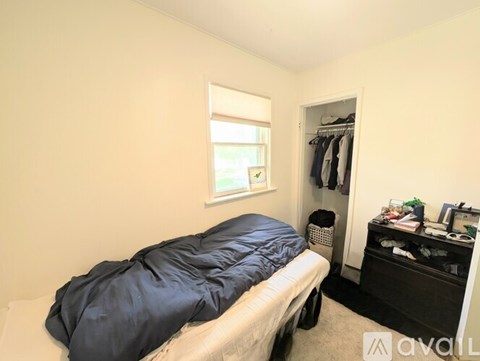 A bedroom with a bed covered in a blue sleeping bag, a window with a view of greenery outside, and a wardrobe with clothes hanging on it.