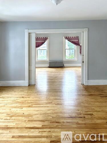 A room with wooden floors and white walls, leading to a hallway with a window.