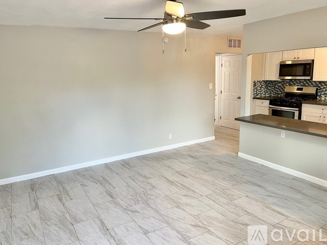 A room with a fan and a kitchen area in the background.