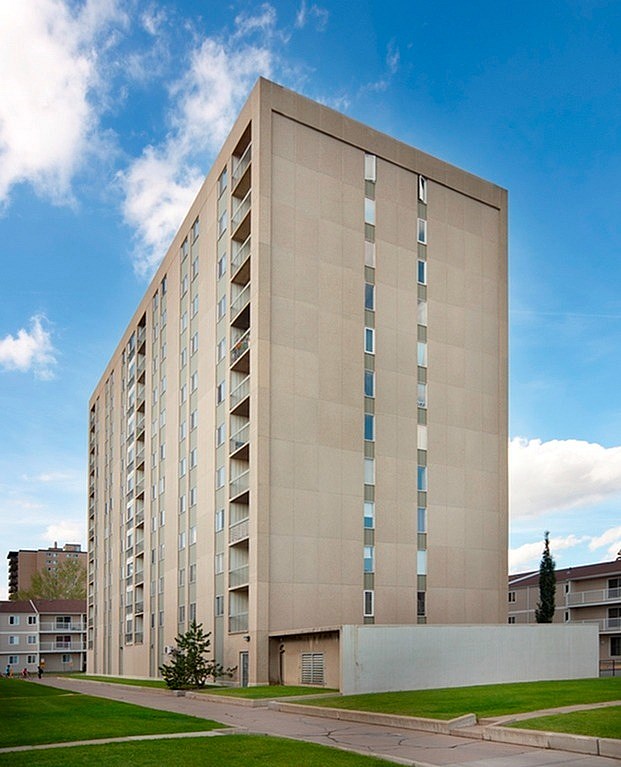 A tall beige building with many windows is surrounded by a green lawn and other buildings.