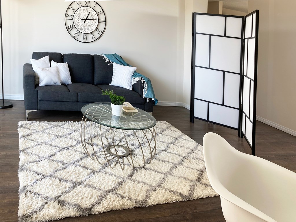 A living room with a grey sofa, a glass table, a white chair, a black and white clock, and a black folding screen.