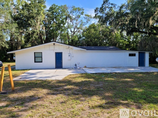 A white building with a black roof and a blue door is surrounded by trees.