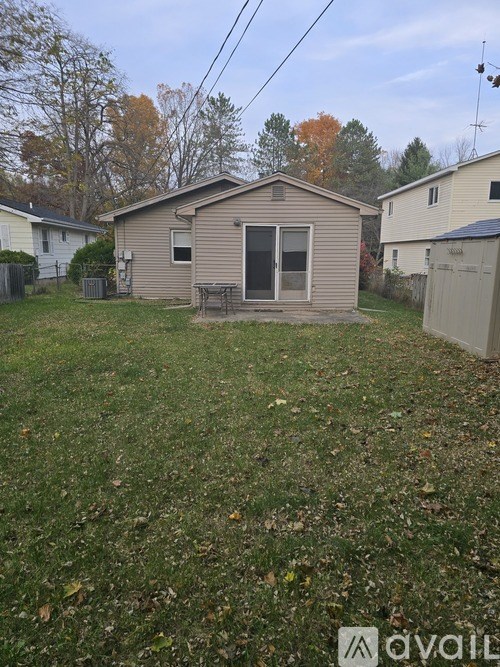 A small house with a brown roof and a grey wall is surrounded by a grassy area with fallen leaves.