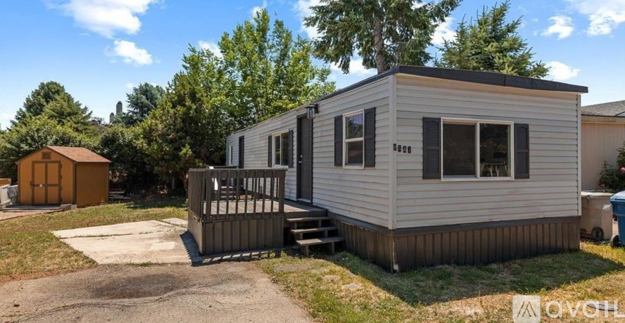 A small house with a brown roof and a grey exterior is surrounded by trees and has a wooden deck.