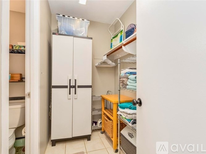 A small, white, two-door refrigerator is in a narrow room with a shelf above it.