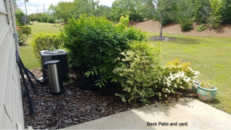 A back patio and yard with a trash can and bushes.
