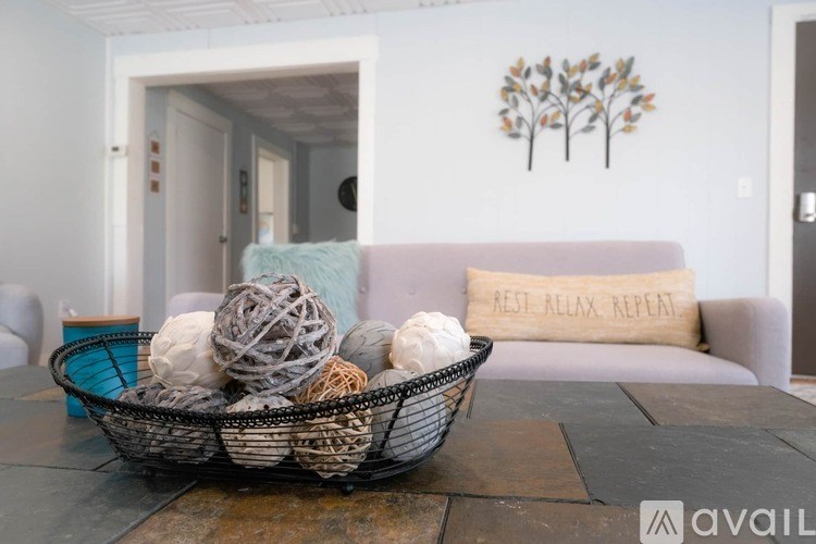 A basket filled with balls of yarn sits on a coffee table in a room with a grey couch and a pillow that reads "Best Allyx Repeat".