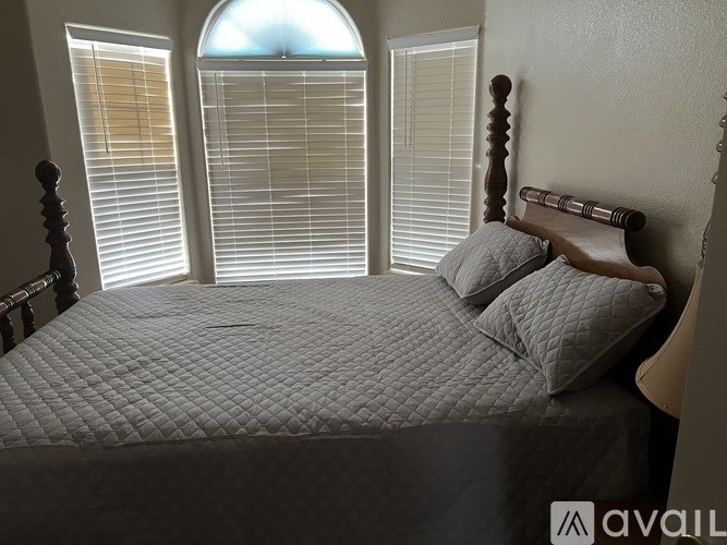 A bed with a wooden headboard and a grey comforter with a quilted pattern is in front of a window with blinds.