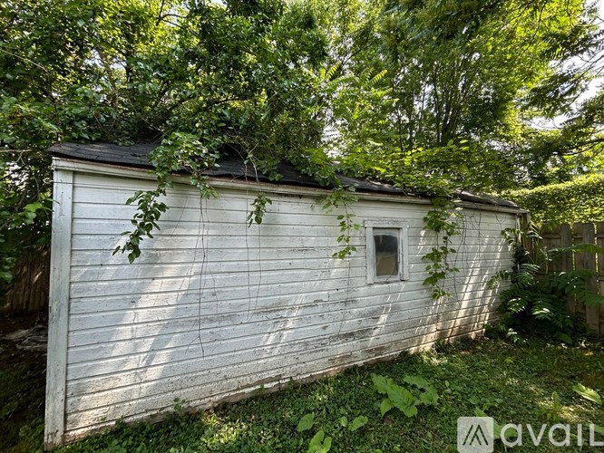 A white wooden shed with a window is surrounded by greenery.