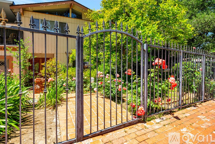 A metal fence with a gate is in front of a house with a brick walkway and red flowers.