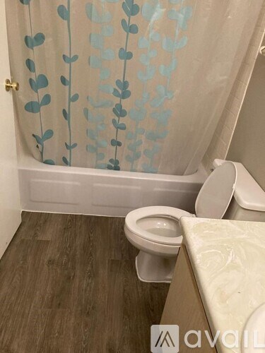 A bathroom with a toilet, a shower curtain with a leaf pattern, and wooden flooring.