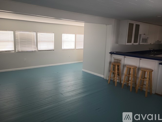 A room with wooden floors and white walls, featuring a kitchen area with bar stools.