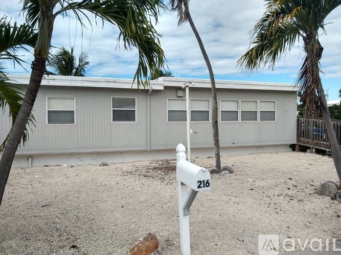 A white mailbox with the number 216 on it is in front of a building with a fence and palm trees.