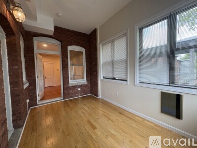 A room with wooden floors and brick walls has a window with blinds and a door leading to another room.