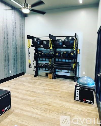 A gym room with a wooden floor, a wall-mounted rack with black weight plates, a fan, and a black device on the floor.