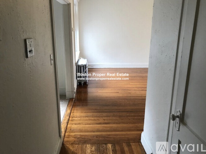 A room with wooden flooring and white walls, with a door on the left and a wall-mounted light fixture.