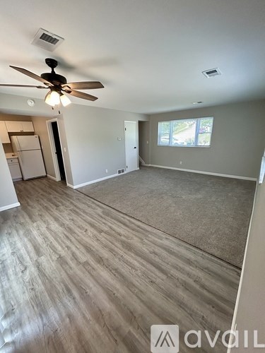 A room with a ceiling fan and light fixture, and a kitchen area with a refrigerator in the background.