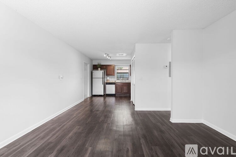 A spacious room with wooden floors and white walls, leading to a kitchen area.