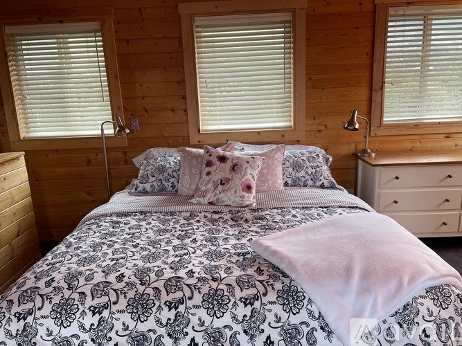 A bed with a black and white patterned comforter and pink pillows sits in a room with wooden walls and windows.