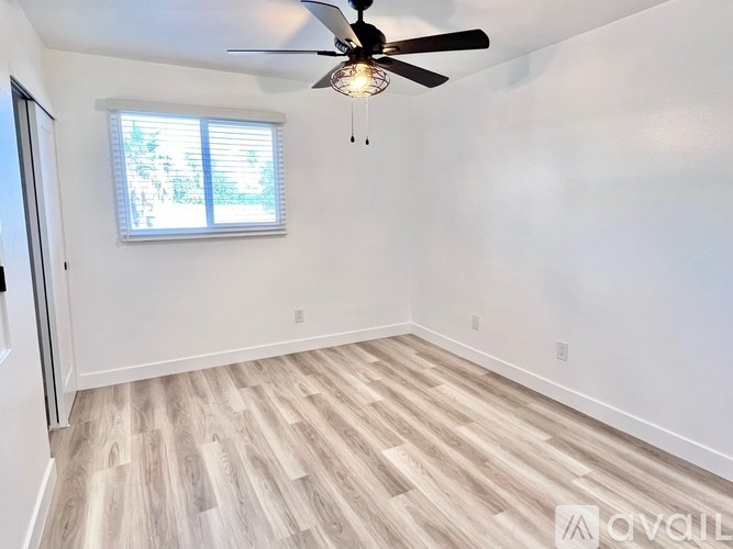 A room with a ceiling fan and light, wooden flooring, and a window with blinds.