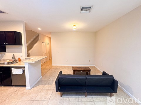 A living room with a black couch and a kitchen area with a sink and microwave.