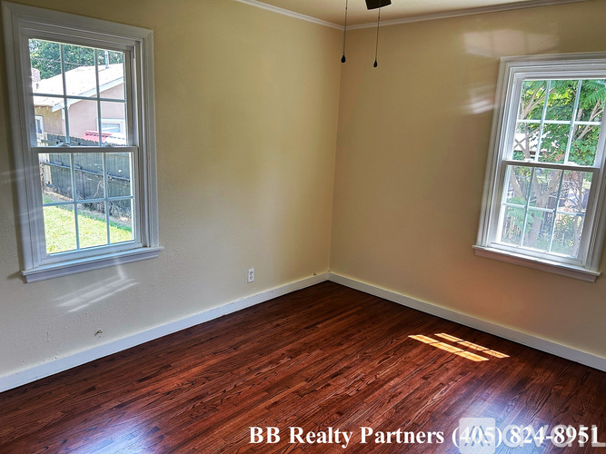 A room with wooden floors and two windows, one of which has a view of a fence and trees.