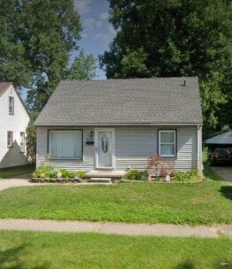 A small house with a grey roof and white siding is surrounded by greenery.