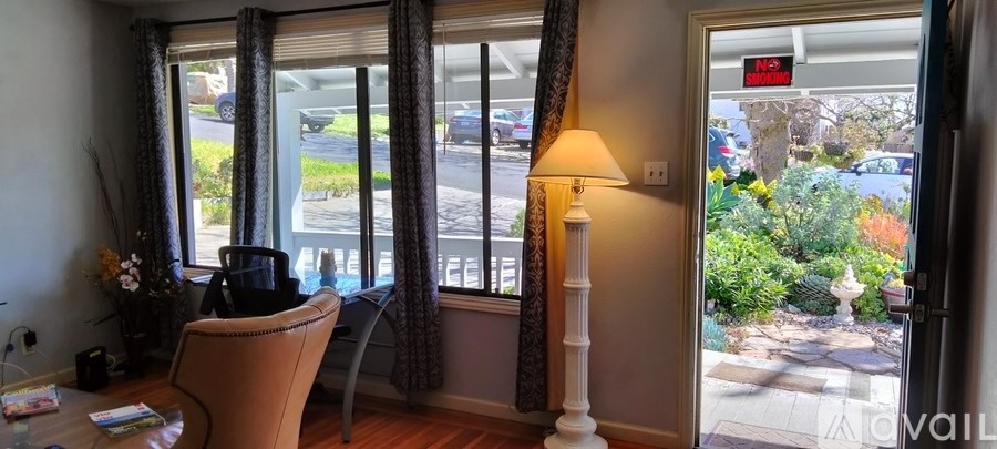A room with a chair, a table, a lamp and a window with a view of the street.