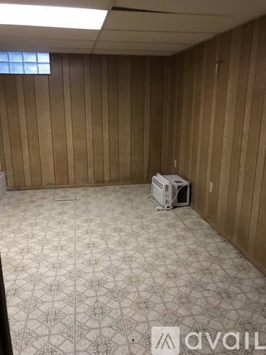 A room with wooden walls and a patterned carpet has a window with blinds and a computer tower on the floor.