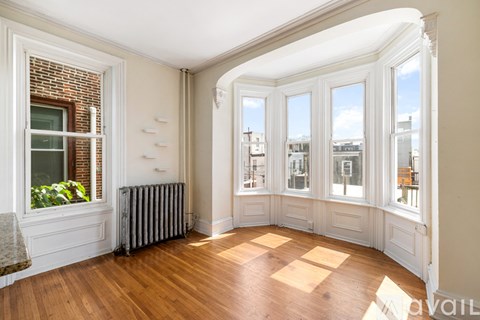 A room with wood floors and white walls has a window with a view of trees.