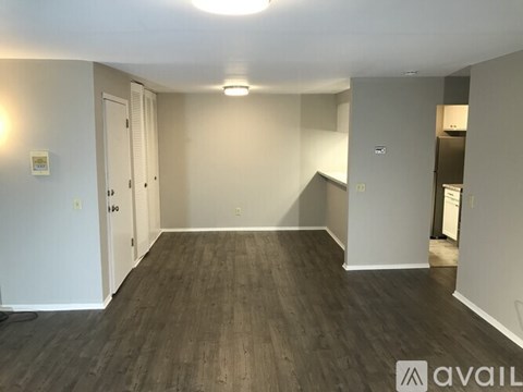 A room with wooden flooring and white walls, with a door on the left and a kitchenette on the right.