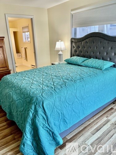 A bed with a teal comforter and two matching pillows sits in a room with a wooden floor and a black headboard.