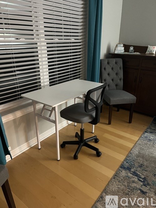 A white desk with a black chair in front of a window with blinds.