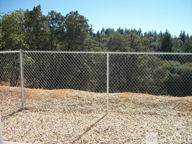 A chain link fence in front of a gravel area with trees in the background.
