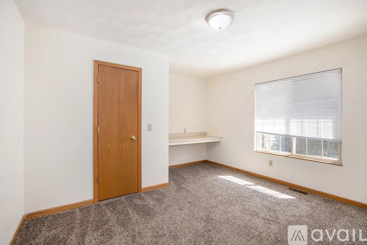 A room with a carpeted floor, a wooden door, a window with blinds, and a white wall.