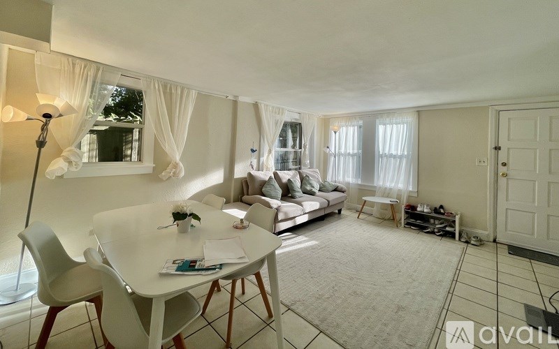 A living room with a white dining table and chairs, a sofa, and a large window with curtains.