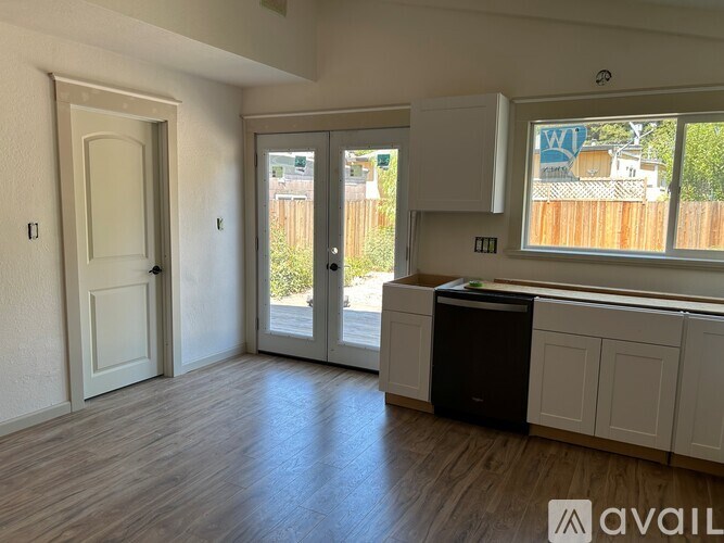 A room with a white door, a window with a view of a fence and houses, and a black trash bin on the floor.